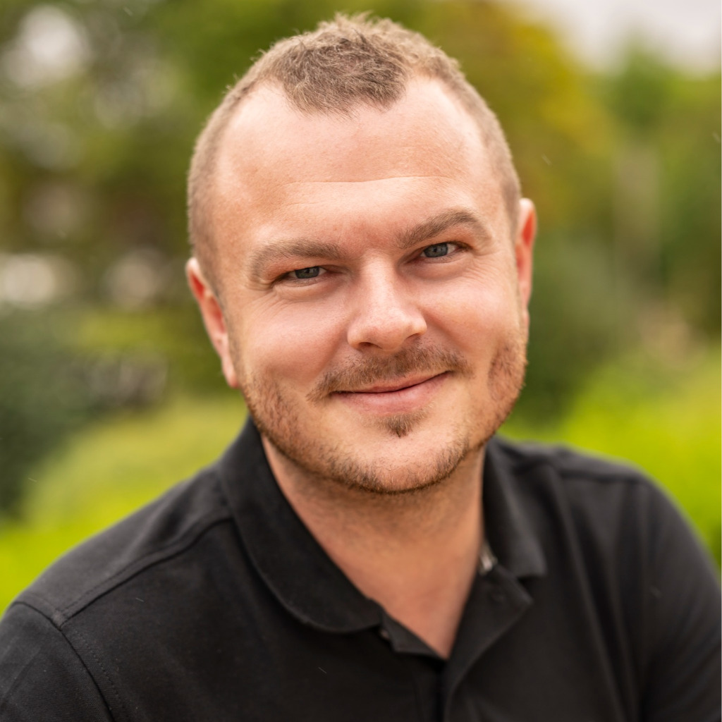 A man with short hair wearing a black polo shirt smiles at the camera with a blurred green background.