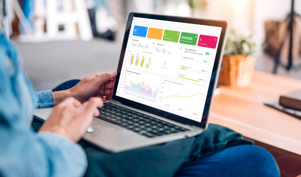 Person using a laptop with a colourful analytics dashboard on the screen, sitting at a desk with a plant in the background.