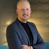A man in a blue blazer and black turtleneck smiles at the camera with his arms crossed. Professional and approachable, he exudes confidence and friendliness.