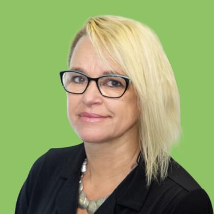 A woman with blonde hair and glasses smiling at the camera, dressed in a black blazer and a necklace against a green background.