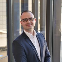 A man in a blue suit standing in front of a glass wall, smiling slightly. He appears to be in a professional setting.