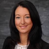 A woman smiling warmly at the camera, dressed in professional attire against a gray background.