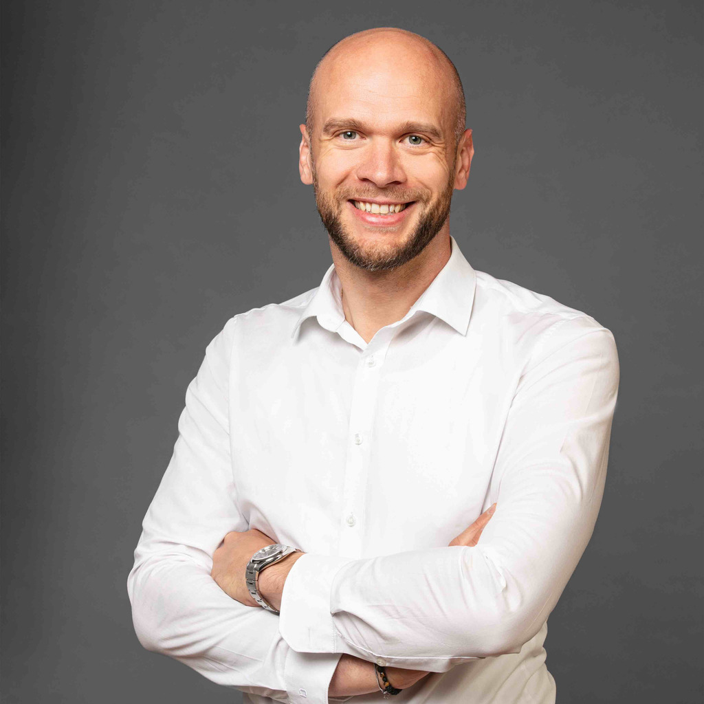 A smiling bald man with a beard, wearing a white shirt and a watch, standing with his arms crossed.