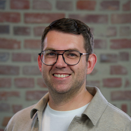 A smiling man wearing glasses and a casual outfit stands in front of a brick wall, exuding a friendly and approachable demeanor.