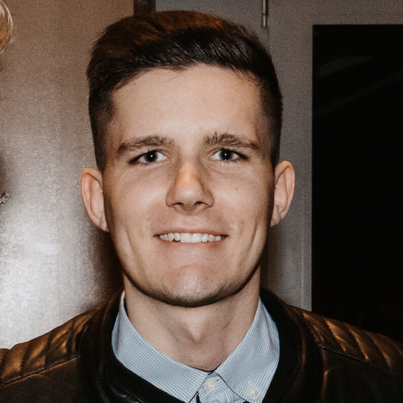 A smiling young man with short brown hair wearing a black leather jacket over a light blue collared shirt.